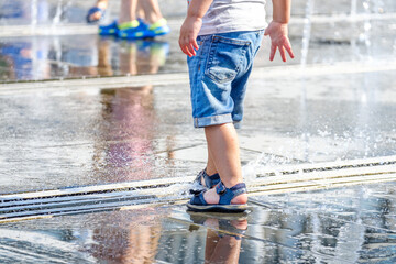 The feet of a child in sandals play with the jets of the fountain in the square. Selective focus. The city is very hot.