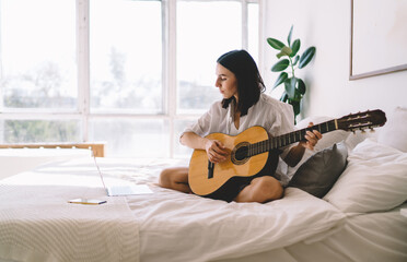 Brunette female learning how to play guitar while watching online video course on laptop computer in cozy home interior. Young woman with netbook enjoying hobby with musical instrument relaxing on bed