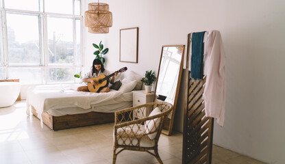 Brunette female learning how to play guitar while watching online video course on laptop computer in cozy home interior. Young woman with netbook enjoying hobby with musical instrument relaxing on bed