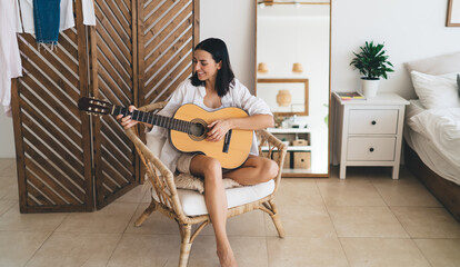 Brunette attractive female smiling and learning to play guitar while sitting in cozy home interior. Young happy woman enjoying her hobby with musical instrument performance relaxing on wicker chair