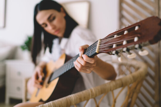 Selective Focus Effect, Female Learning To Play Guitar While Sitting In Cozy Home Interior. Young Concentrated Woman Enjoying Her Hobby With Musical Instrument Performance Relaxing On Wicker Chair