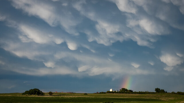 Storm clouds above farmland with a small rainbow peaking out below the fluffy Mammatus clouds