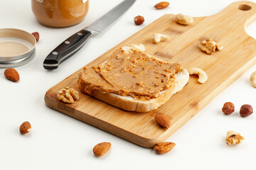 Square bread for toast with peanut butter on a wooden board. Nuts, a knife, a jar of peanut butter, and a wooden cutting board with a sandwich on a white table.