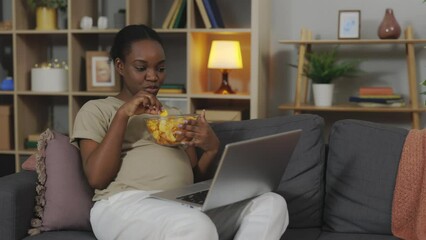 African american expectant mother having unhealthy snack while watching favorite program on laptop. Attractive pregnant woman relaxing alone at home with pleasure.