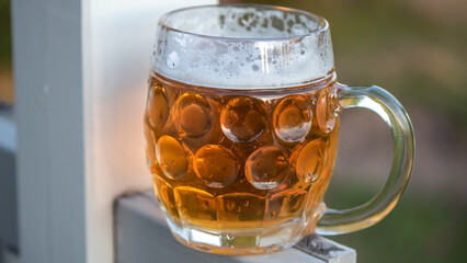 Sun sets behind recently poured lager beer. The beverage rests in an ovally shaped beer glass with a handle. The gold and amber color of the beer makes a brilliant contrast to the white foam up top.