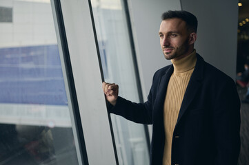 Handsome thoughtful traveler man of Middle-Eastern ethnicity, standing near panoramic windows overlooking runway in airport departure terminal