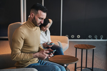 Multitasking Caucasian man using mobile phone, sitting next to his female partner in the VIP lounge of an international airport departure terminal