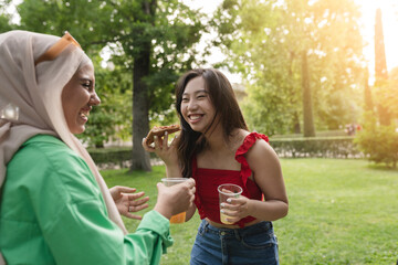 Obraz premium multiracial female friends eat pizza and smile in the park, focus on asian woman.