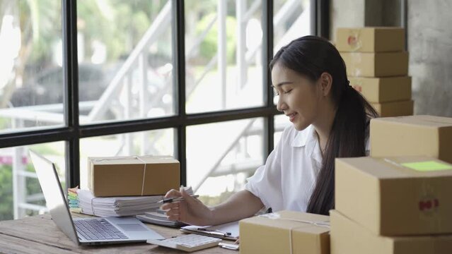 Start an SME, a small business entrepreneur of independent Asian women using laptop with box. cheerful success Woman working on online marketing packaging boxes and delivering SME concepts 4k video