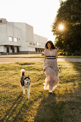 Naklejka na ściany i meble Georgian woman walking with siberian husky at sunset. Attractive girl with her lovely dog playing on the street.