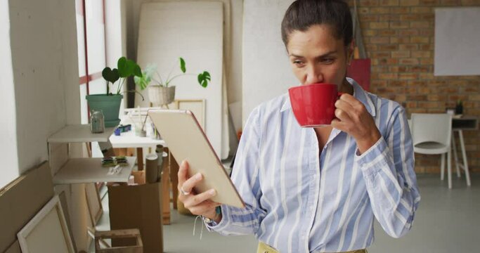 Video of happy biracial female designer using tablet and drinking coffee in studio