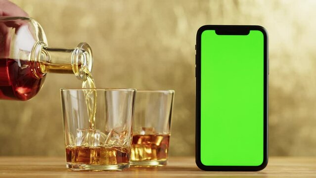 Brandy or whiskey close-up. Luxury cognac and smartphone with green chroma key screen. Alcohol amber drink with ice, drinking rum, liqour beverage in glass.