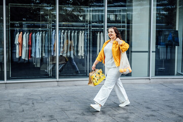 Happy confident smiling plus size curvy young woman with shopping bags walking on city street near shop windows