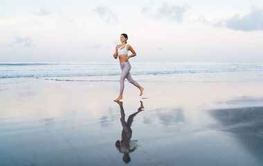 Caucasian woman in stylish tracksuit running at seashore for keeping perfect slim figure, barefoot energetic female in sportive clothes have cardio training while jogging at coastline beach
