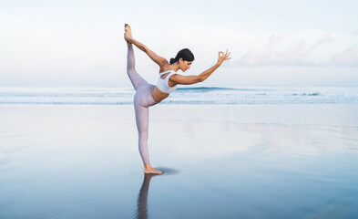 Fototapeta premium Caucaisan woman with good body shape concentrated on hatha asana pose with raised leg, young female in sportive tracksuit stretching for feeling vitality during holistic healing on seashore retreat