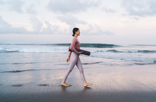 Side view of slim woman in sportive clothes holding mat and walking near calm sea waves spending weekend morning for training at nature, charming female in tracksuit choosing place for stretching