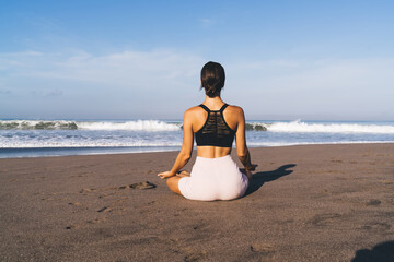 Fototapeta premium Back view of female yogi thinking about zen mindfulness sitting in lotus at coastline beach, woman in tracksuit relaxing during morning workout for searching harmony during meditation practice