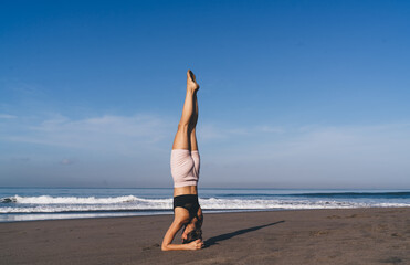 Fototapeta na wymiar Slim female athlete doing headstand exercise during morning workout near ocean and natural horizon,Caucaisan fit girl practice strength and body balance keeping healthy lifestyle and reaching vitality