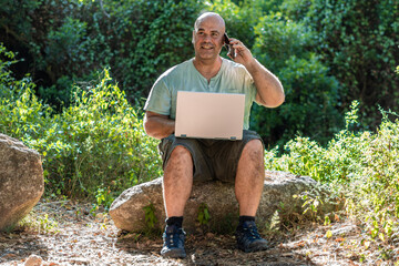 Sweaty man sitting on a rock in the forest, with a laptop on his knees and talking on the phone.