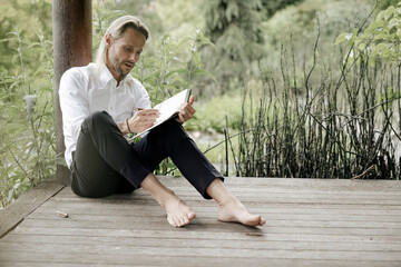 Geschäftsmann im weißen Hemd sitzt auf Holzsteg und schreibt in ein Buch
