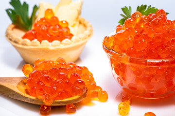 Delicious red caviar in wooden brown spoon, in small crystal cup with top decorated with parsley and red caviar in tartlet with butter and parsley on white background.