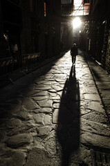 Silhouette of a person walking through a cobbled street receiving direct sunlight and leaving...