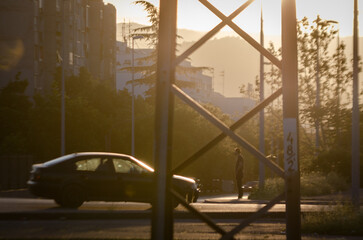 Sunset in a city with trees and residential buildings, while a young man waits standing in front of a car