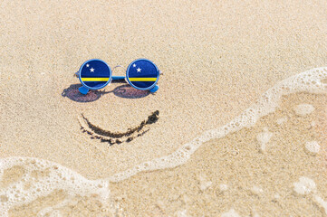 Fototapeta na wymiar A painted smile on the sand and sunglasses with the flag of the Curacao. The concept of a positive and successful holiday in the resort of the Curacao.