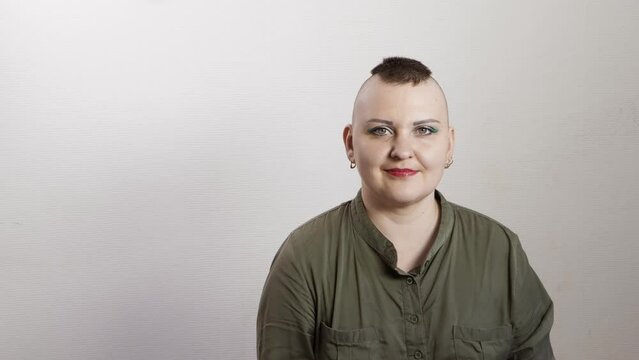 A shaved woman with a mohawk sits on a gray background smiling. Medium plan