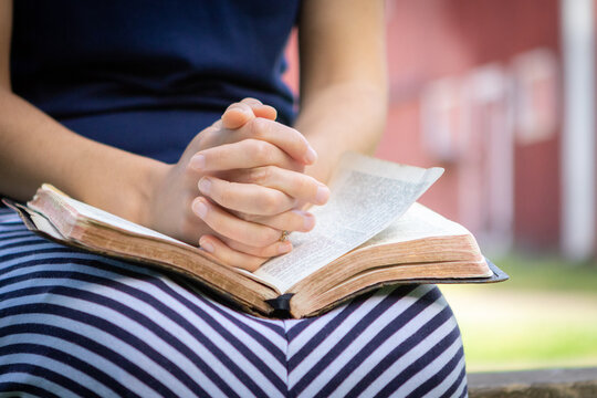 Young Christian woman sitting outside having quiet time with folded hands praying with open Bible in lap