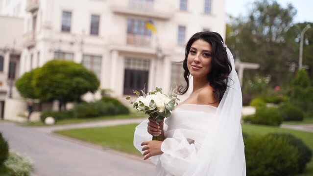 Pretty bride in long airy veil, elegant white dress smells flowers on wedding day. Attractive woman with bouquet looking at camera in slow motion. Fabric fluttering in the air.