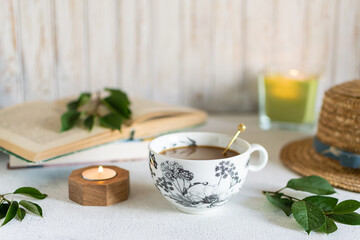 Cozy composition with cup of coffee with milk, interesting book, candles and green leaves on white wooden background. Hygge style composition.