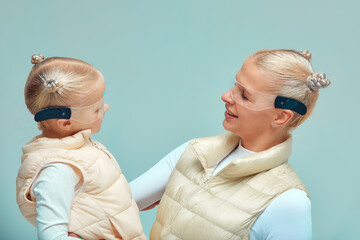 Virtual technology. Happy delighted nice girl smiling and looking to her mother while mother holding in her arms wearing digital virtual glasses on biege background