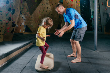 Father supporting daughter training on balance board in a gym