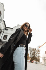 Stylish beautiful young woman in a fashionable black leather long coat with high-waisted jeans and a tank top is fixing cool sunglasses in the city. Gorgeous female urban style dress