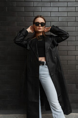 Stylish glamorous young pretty woman model in fashionable black clothes with a fashion long leather coat with a black T-shirt and vintage jeans standing near a black brick wall