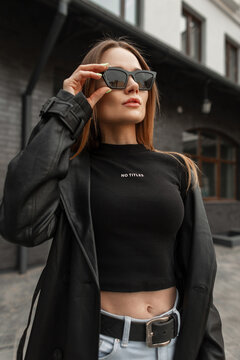 Stylish beautiful young woman model in fashionable black clothes with a black leather coat with a black mock up T-shirt wears vintage cool sunglasses in the city