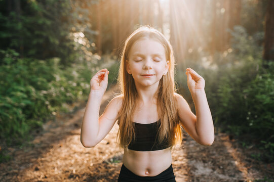 Portrait of hardworking blonde preschool girl, professional athlete child in black suit, sitting cross-legged on green rug in lotus position and meditating practicing yoga exercises at sunset.
