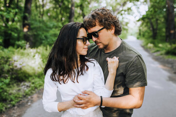 Bearded stylish man and hippie brunette woman in sunglasses hugging and smiling outdoors in the park. Portrait, photograph of people in love.