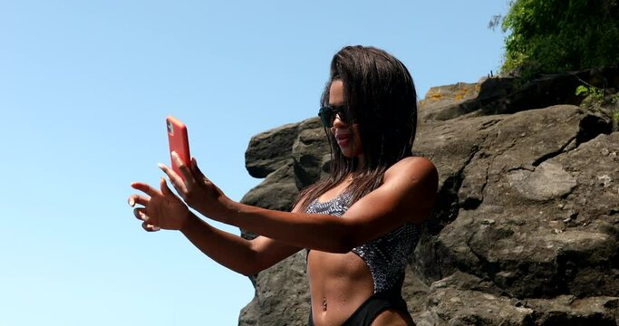 Beautiful black woman with blowing hair using smartphone and making selfie on sea background. 