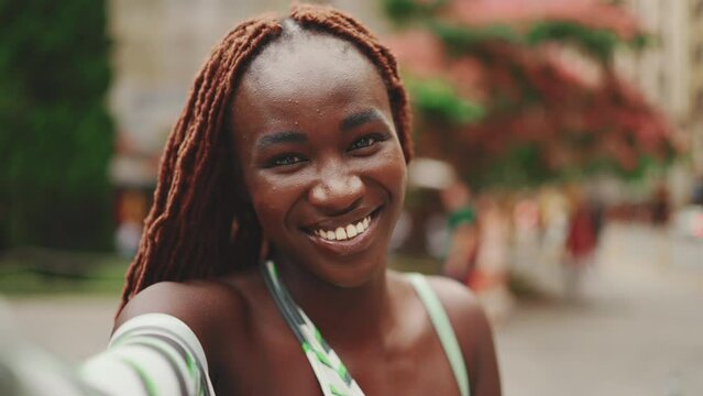 Clouse-up, smiling gorgeous woman with African braids wearing top stands outside on the street and uses mobile phone. Stylish girl taking selfie photos