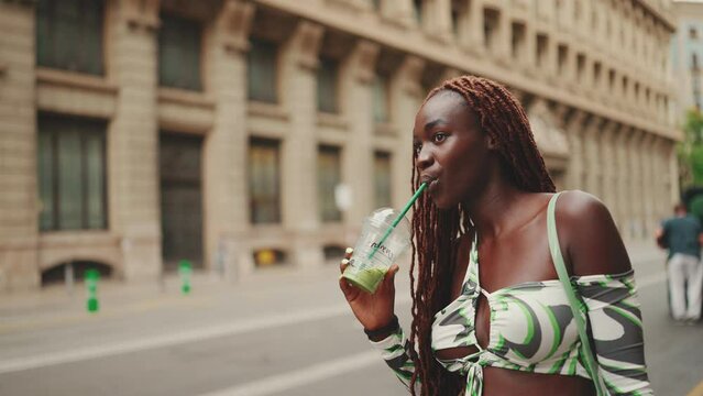 Gorgeous woman with African braids wearing top, with fresh cocktail drink in plastic cup in her hands, standing on the sidewalk next to the road waiting for taxi