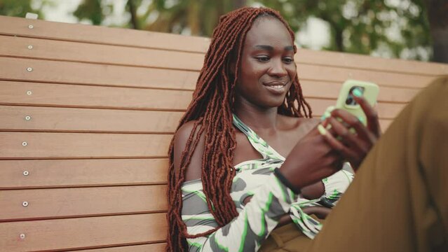 Smiling gorgeous woman with African braids wearing in top is using mobile phone while sitting on bench. Stylish girl watching photo, video on her smartphone