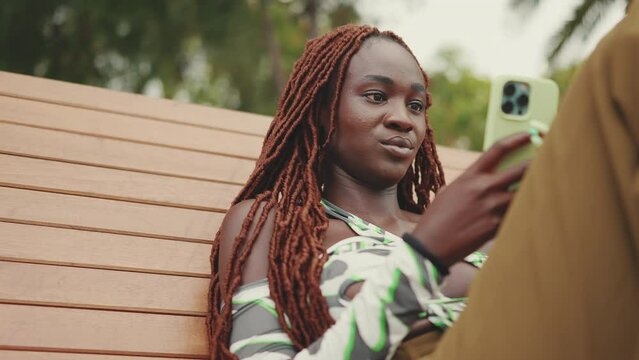 Gorgeous woman with African braids wearing in top is using mobile phone while sitting on bench. Stylish girl watching photo, video on her smartphone. Camera zooms