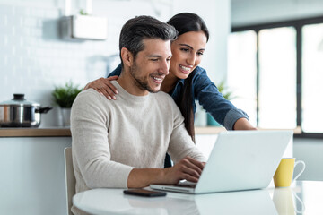 Beautiful lovely couple using their laptop while having breakfast in the kitchen at home.