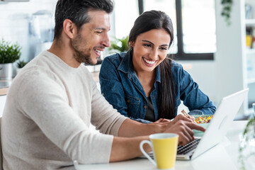Beautiful lovely couple using their laptop to searching voyage while eating poke bowl in the kitchen at home.