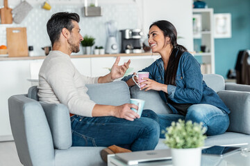 Lovely couple holding coffee cups while talking sitting on couch at home.