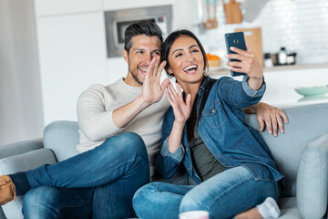 Naklejka na ściany i meble Happy young couple making a video call with smart phone while sitting on couch at home.