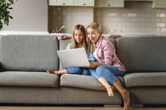 Friendship of mom and daughter. Cheerful caucasian mom and daughter are sitting on the couch, using a laptop, laughing, browsing the Internet, shopping online, excited of a good news