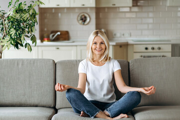 Smiling caucasian middle aged female sits on sofa at living room and  practise yoga, meditation in lotus position looks at camera and smiling, relieve stress